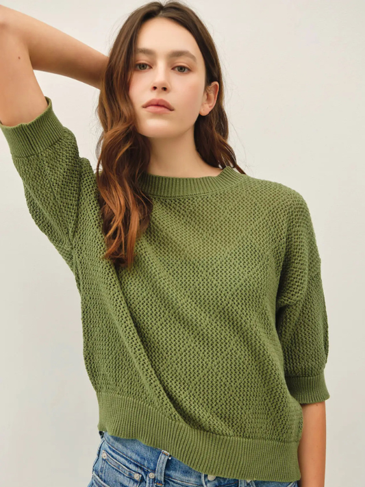 A woman modeling a textured olive green knit sweater with three-quarter sleeves, paired with light-wash blue jeans, against a plain off-white studio background.