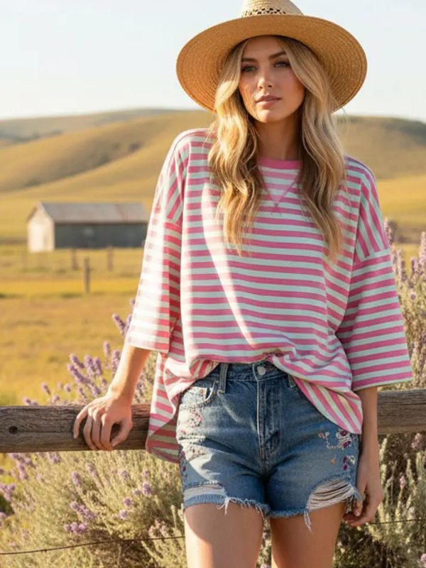 Woman wearing a pink and white striped shirt and denim shorts standing in a field with lavender and a barn in the background.