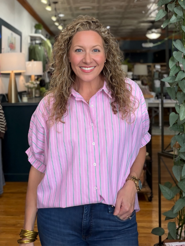 A smiling woman with curly blonde hair wearing a pink and white vertically striped button-down shirt and dark blue jeans, standing in a retail store interior.