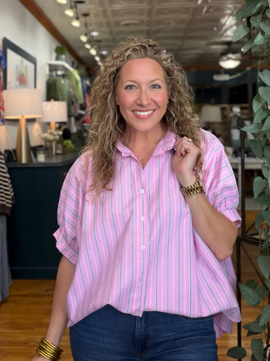 A smiling woman with curly blonde hair wearing a pink and white vertically striped short-sleeve button-up shirt and dark blue jeans, standing inside a retail store.