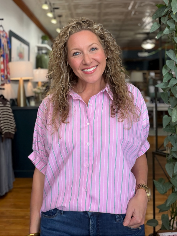 A smiling woman with curly blonde hair wearing a pink and white vertically striped short-sleeve button-down shirt and blue jeans, standing inside a retail store.