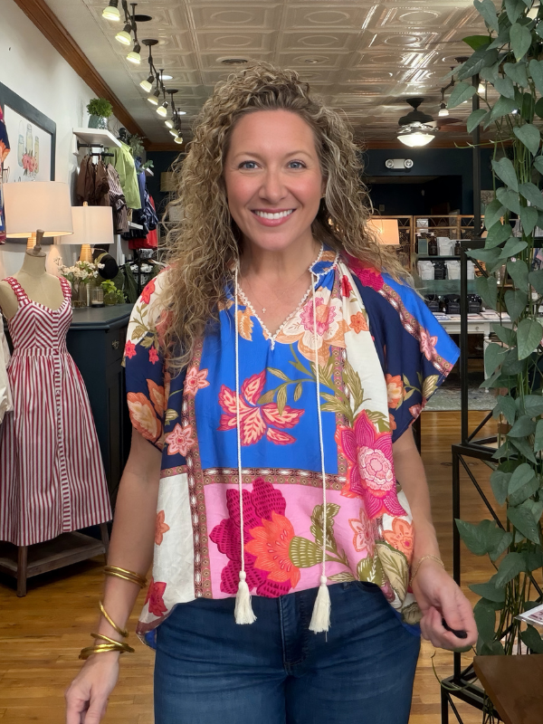 A smiling woman with curly hair stands in a boutique wearing a colorful patchwork floral blouse with tassel ties and blue jeans.