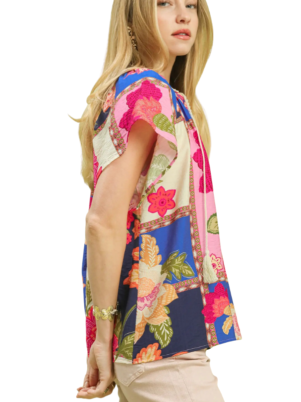 A woman wearing a short-sleeve patchwork floral top with vibrant pink, blue, green, and beige panels, paired with beige pants, shown against a plain white background.