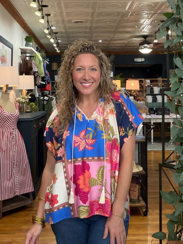 A smiling woman with curly hair stands in a boutique wearing a colorful floral short-sleeve top with blue, pink, orange, and cream sections, paired with dark jeans.
