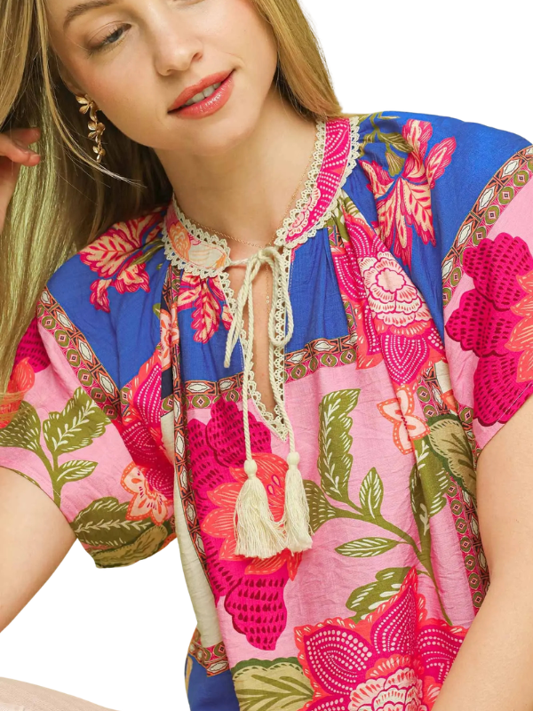 A close-up of a woman wearing a colorful short-sleeve blouse with a pink and blue floral print, lace neckline, and cream tassel tie.