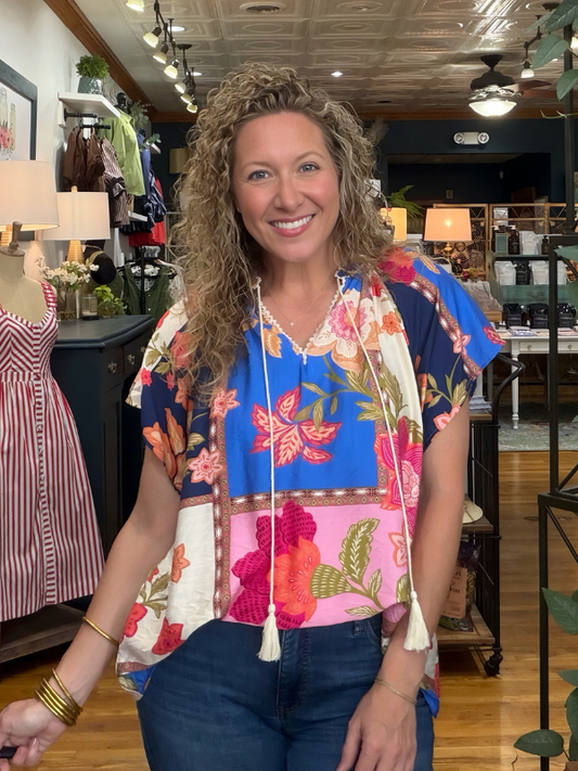 A smiling woman with curly blonde hair stands in a boutique wearing a colorful patchwork floral top with blue, pink, and cream sections, paired with dark blue jeans.