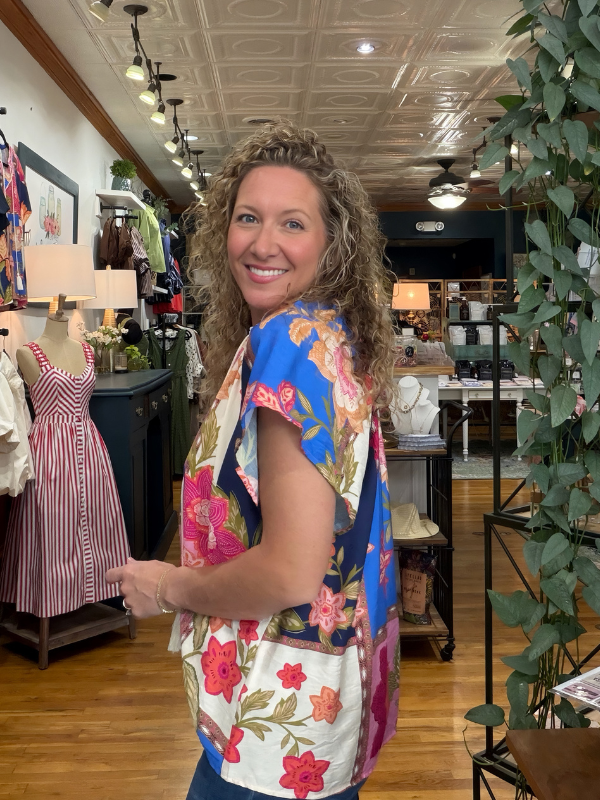 A woman with curly brown hair smiling and looking back over her shoulder inside a boutique, wearing a colorful floral print short-sleeve top with blue, pink, white, and green tones.