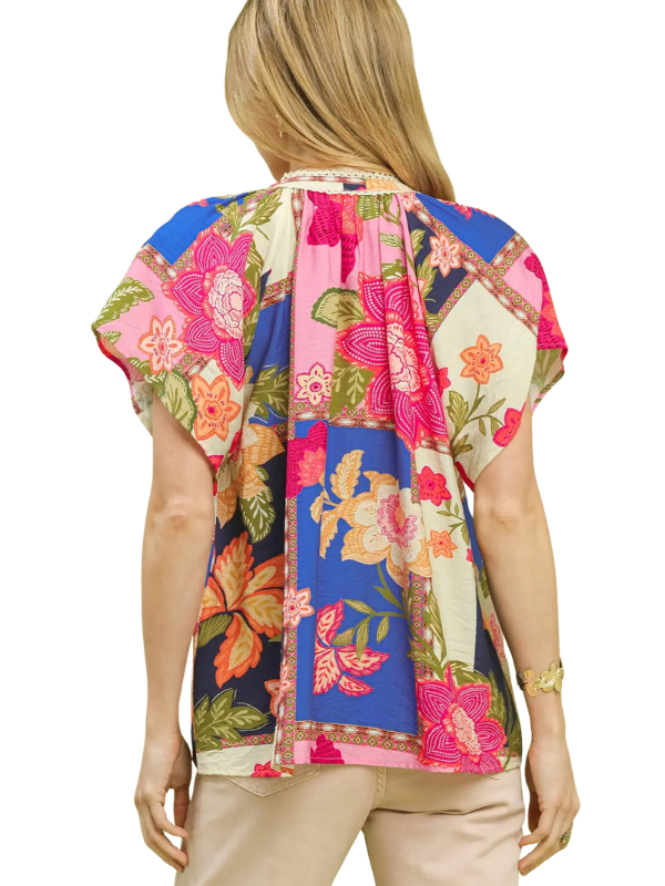 A woman shown from the back wearing a short-sleeve patchwork blouse with a vibrant floral print in pink, blue, green, and beige, paired with beige pants against a white background.