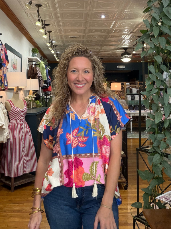A smiling woman with curly hair stands inside a boutique wearing a colorful floral short-sleeve top with blue, pink, and white patterns, paired with dark blue jeans.