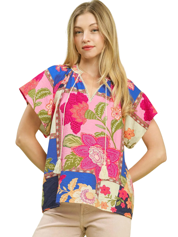 A woman modeling a short-sleeve V-neck top with a colorful patchwork floral print, featuring pink, blue, green, and beige tones, set against a plain white background.
