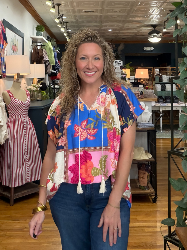 A smiling woman with curly hair stands in a boutique shop wearing a colorful short-sleeve top with a vibrant floral print, blue jeans, and gold bracelets.