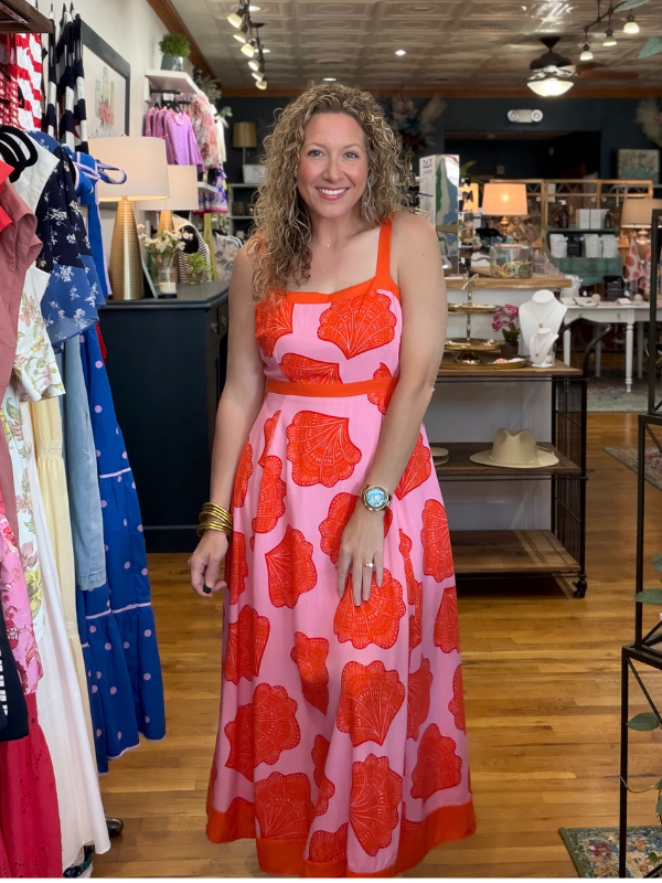 Woman in a pink and red floral dress standing in a store with clothing racks and displays.