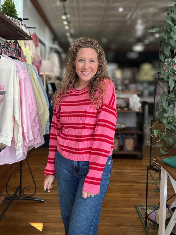 Woman wearing a pink striped sweater and blue jeans standing in a store with clothing racks and shelves in the background.