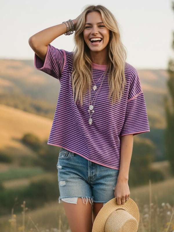 Woman wearing a purple striped shirt and denim shorts standing in a field with mountains in the background.
