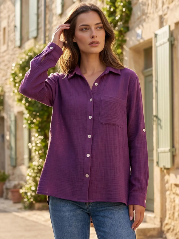 A woman wearing a textured purple long-sleeve button-up shirt and blue jeans, standing outdoors on a sunlit stone street with green shutters and foliage in the background.