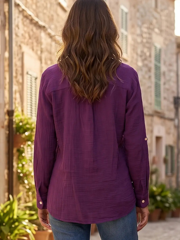 A woman viewed from behind wearing a textured purple long-sleeve button-up shirt and blue jeans, standing outdoors in front of rustic stone buildings.