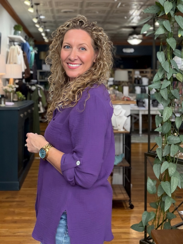 A smiling woman with curly blonde hair stands in a retail store wearing a purple button-up shirt with rolled sleeves and denim shorts.
