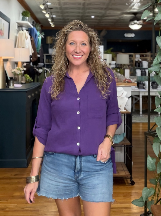 A smiling woman with curly blonde hair stands in a retail store wearing a purple button-up shirt and denim shorts.