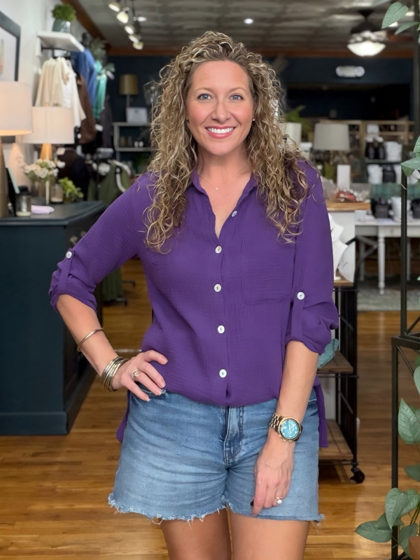A smiling woman with curly blonde hair stands in a retail store wearing a purple button-up shirt and denim shorts.