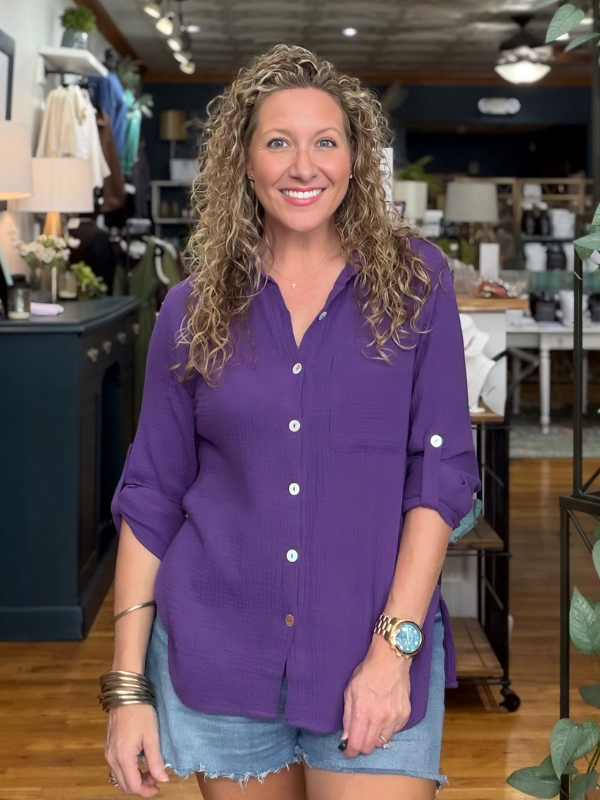 A smiling woman with curly blonde hair stands in a retail store wearing a purple button-up shirt with rolled sleeves and denim shorts.