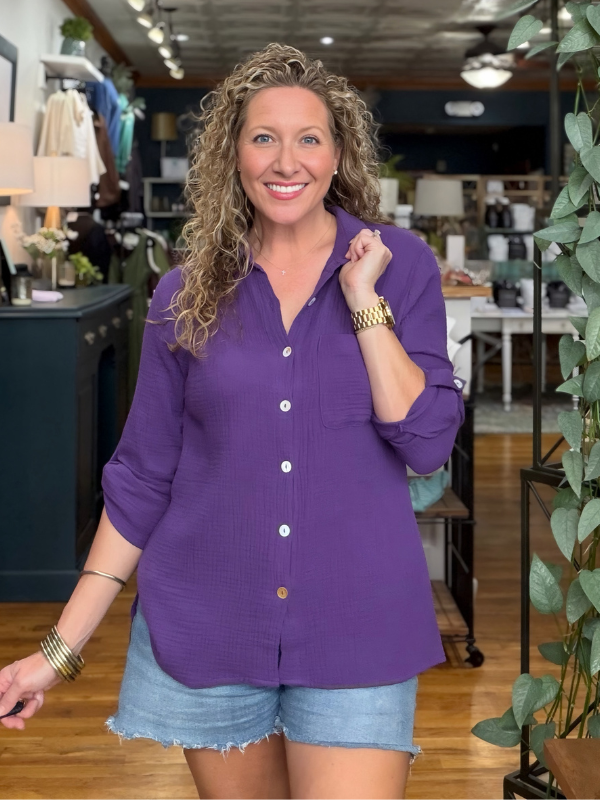 A smiling woman with curly blonde hair wearing a purple button-down shirt and denim shorts, standing inside a retail store.