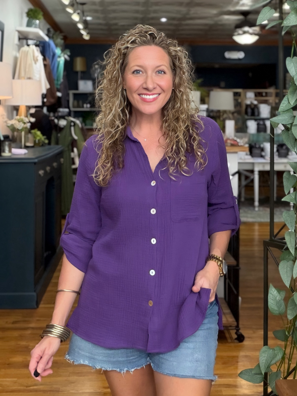 A smiling woman with curly blonde hair stands in a clothing store wearing a purple long-sleeve button-up shirt and denim shorts.