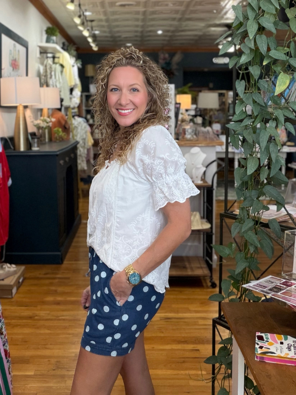 Woman in a white blouse and blue polka dot shorts standing in a store with plants and furniture.
