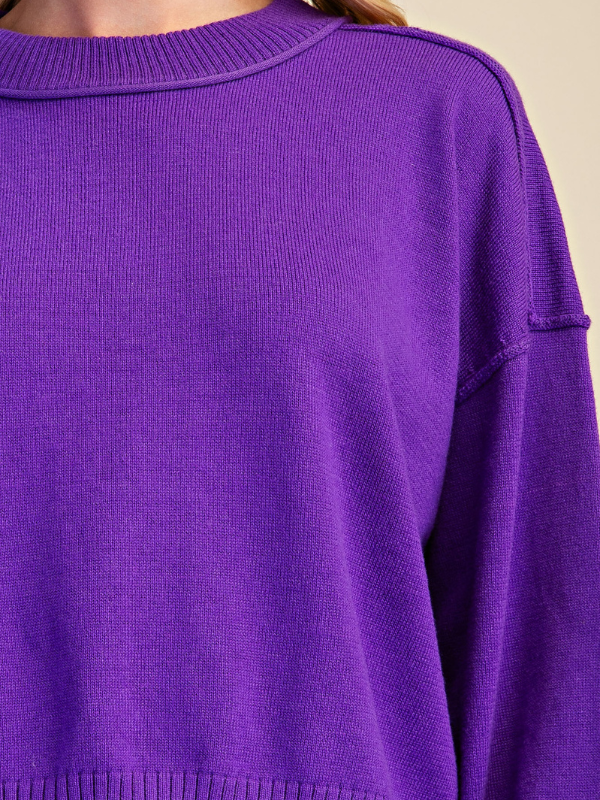 Close-up of a person wearing a vibrant purple ribbed knit sweater, showing the crew neck and shoulder details against a neutral beige background.