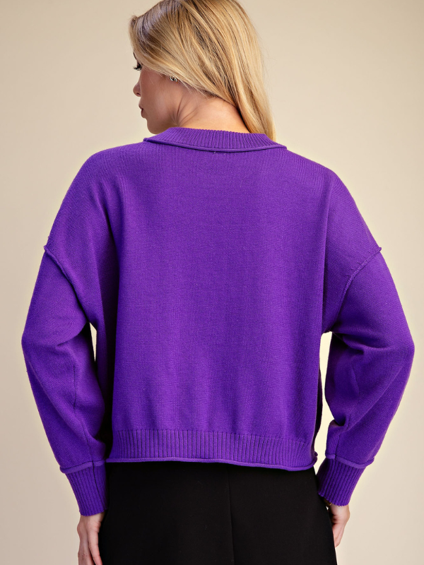 A woman shown from the back wearing a vibrant purple knit sweater with a crew neck and black pants, set against a solid beige background.