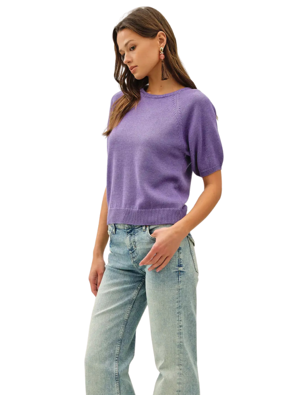 A woman wearing a short-sleeve knit sweater in lavender purple and light-wash blue jeans, standing against a plain white background.