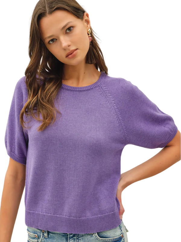 A woman wearing a short-sleeve knit sweater in a soft lavender-purple color, paired with blue jeans, against a plain white background.
