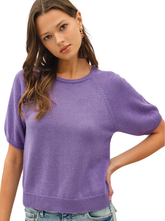 A woman wearing a short-sleeve knit sweater in a soft lavender-purple color, paired with blue jeans, against a plain white background.