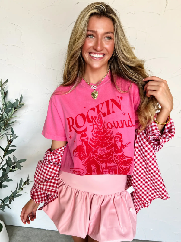 Woman wearing a pink 'Rockin' Around' shirt with a checkered sleeveless top underneath, standing against a white wall.