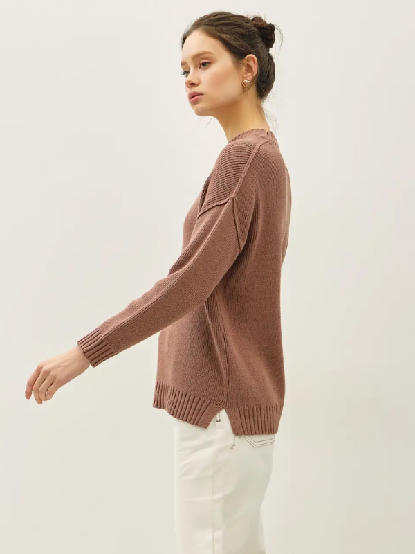 Woman wearing a brown sweater and white pants against a plain background