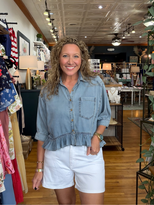 Woman in a denim shirt and white shorts standing in a store with clothing racks and decor.