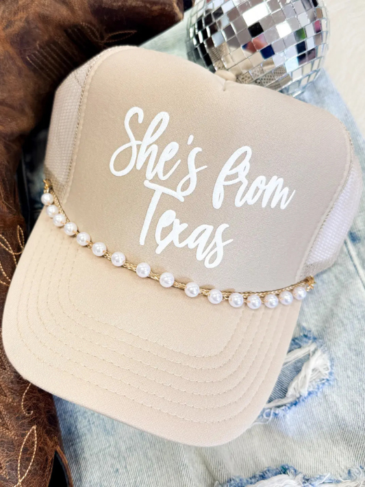 Beige trucker hat with 'She's from Texas' text and pearl embellishments on a denim background.