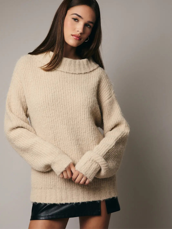 Woman wearing a beige sweater over a black leather skirt against a plain background