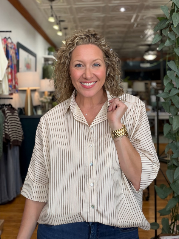 A smiling woman with curly blonde hair wearing a cream and navy blue vertically striped short-sleeve button-down shirt, standing inside a boutique.