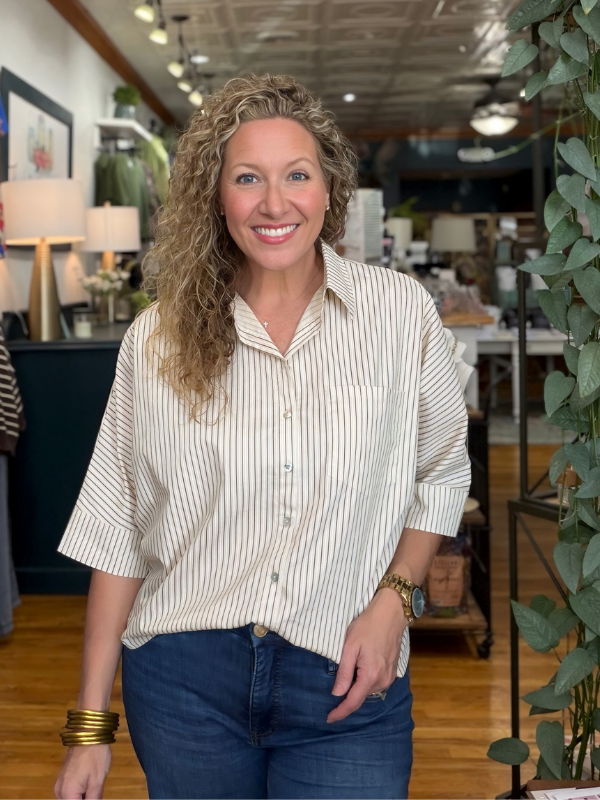 A smiling woman with curly blonde hair stands in a furniture or home decor store wearing a cream and navy vertically striped short-sleeve button-up shirt and dark blue jeans.