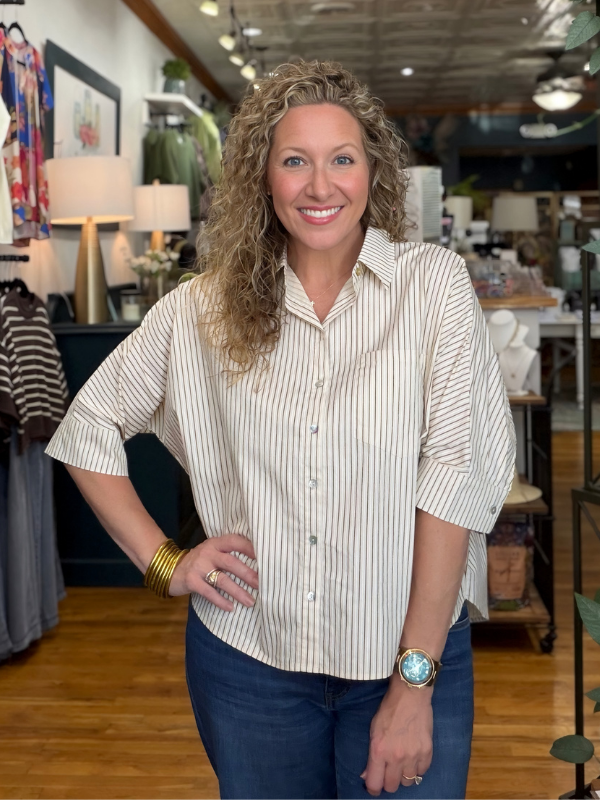A smiling woman with curly blonde hair stands in a boutique wearing a cream and navy vertically striped button-down shirt with three-quarter sleeves and dark blue jeans.