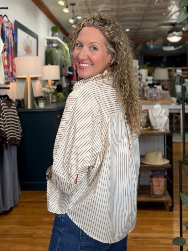 A woman with curly blonde hair wearing a cream and brown vertically striped button-down shirt and blue jeans, posing inside a boutique store.