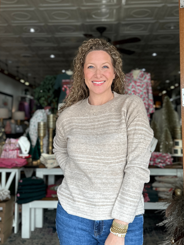 Woman wearing a gray sweater and blue jeans standing in a store with various items in the background.