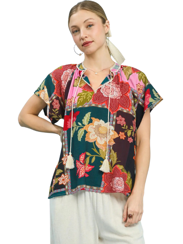 A woman wearing a short-sleeve V-neck blouse with a colorful floral and butterfly print, featuring cream drawstrings with tassels, paired with white pants, against a plain white background.