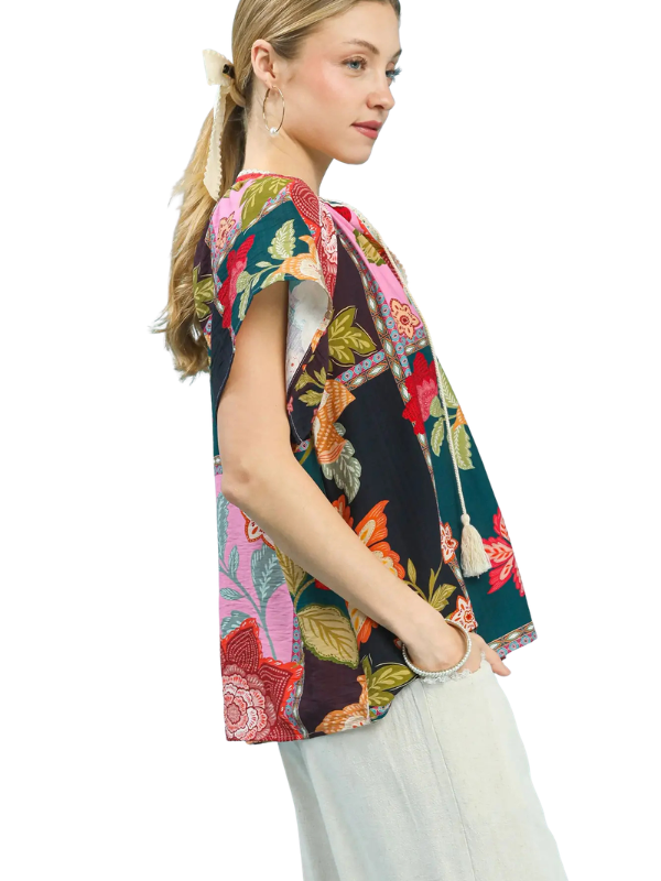A woman wearing a short-sleeve, V-neck top with a vibrant multicolor floral and botanical print, paired with light gray wide-leg pants, shown against a plain white background.