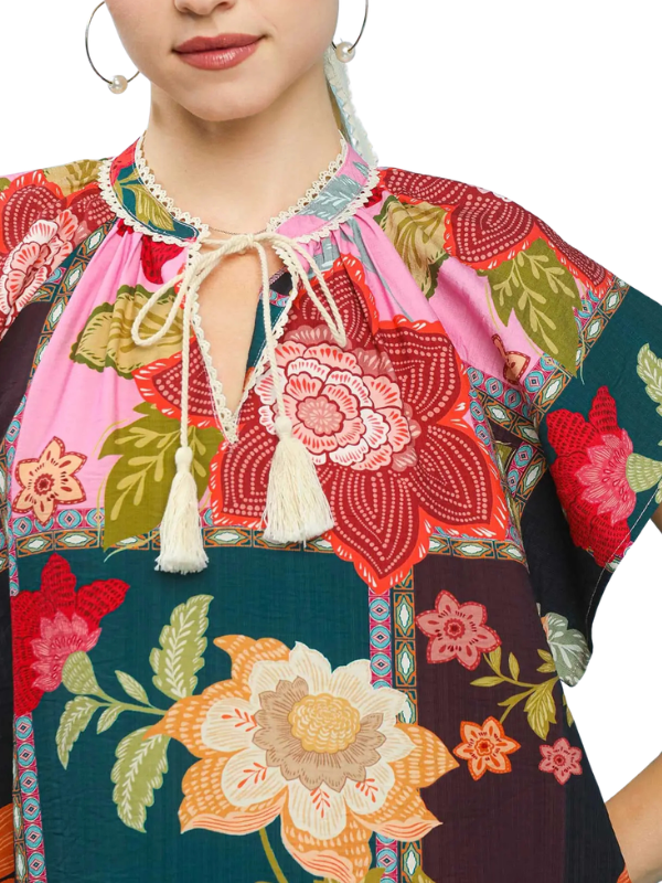 A close-up of a woman wearing a colorful bohemian floral print tunic top with a tie-neck and tassel detail. The top features a vibrant patchwork design with pink, red, green, and dark teal sections.