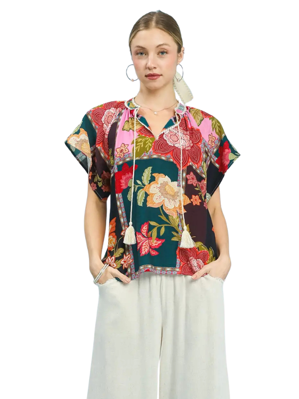 A woman modeling a short-sleeve, V-neck blouse with a colorful floral and paisley print, featuring drawstrings with tassels, paired with light gray wide-leg pants against a plain white background.