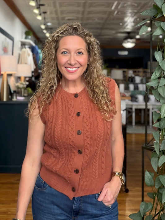 A smiling woman with curly blonde hair stands in a retail store wearing a sleeveless rust-orange cable-knit button-front top and blue jeans.