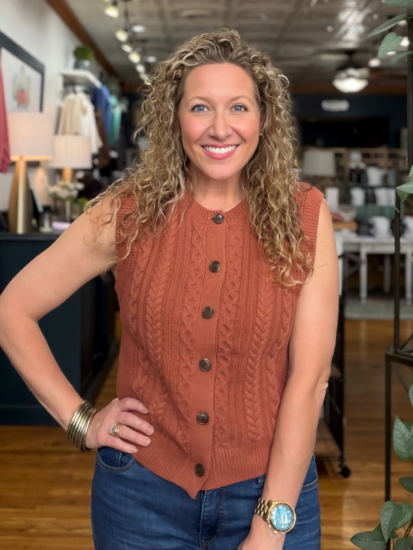 A smiling woman with curly blonde hair wearing a sleeveless rust-orange cable-knit button-front top and blue jeans, standing in a retail store interior.
