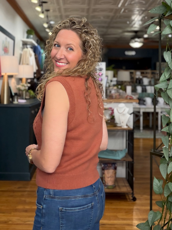 A woman with curly blonde hair wearing a sleeveless rust-orange knit vest and blue jeans, standing in a boutique store.