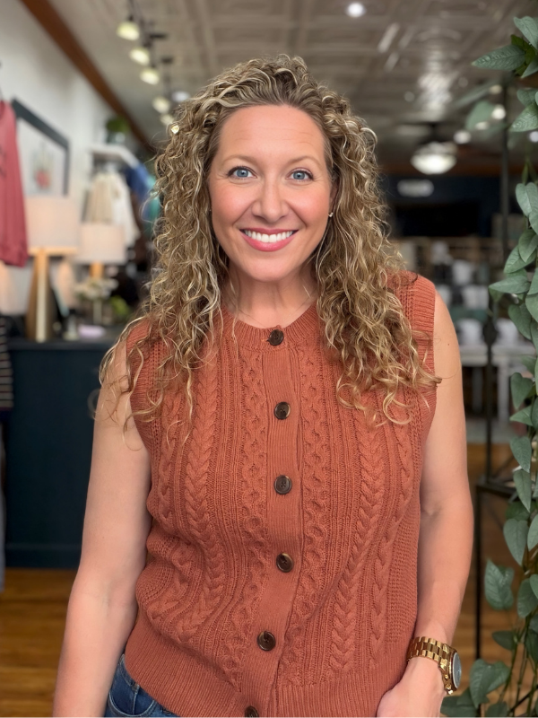 A smiling woman with curly blonde hair wearing a sleeveless rust-orange cable-knit button-front vest, standing in a retail store interior.